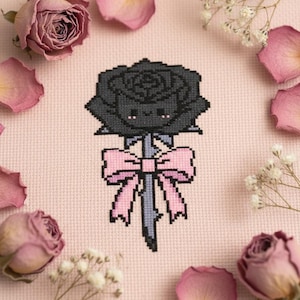 May include: Cross-stitch design of a black rose with a pink bow and stem. The rose has a cute face. The background is a light pink fabric, surrounded by dried roses and baby's breath.