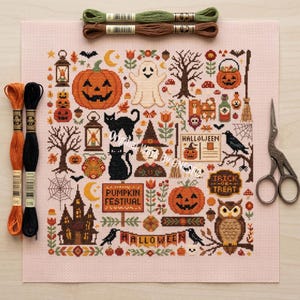 May include: A cross-stitch pattern on light pink fabric featuring Halloween motifs including pumpkins, a ghost, black cats, an owl, a haunted house, and signs reading Pumpkin Festival, Halloween, and Trick or Treat, with embroidery floss and scissors.