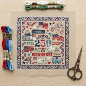 May include: A cross-stitch project with a patriotic theme, featuring American flags, fireworks, and the text "America" and "Independence." The design is framed with a blue and red border. Embroidery floss in red, blue, green, and white, along with scissors, are also visible.