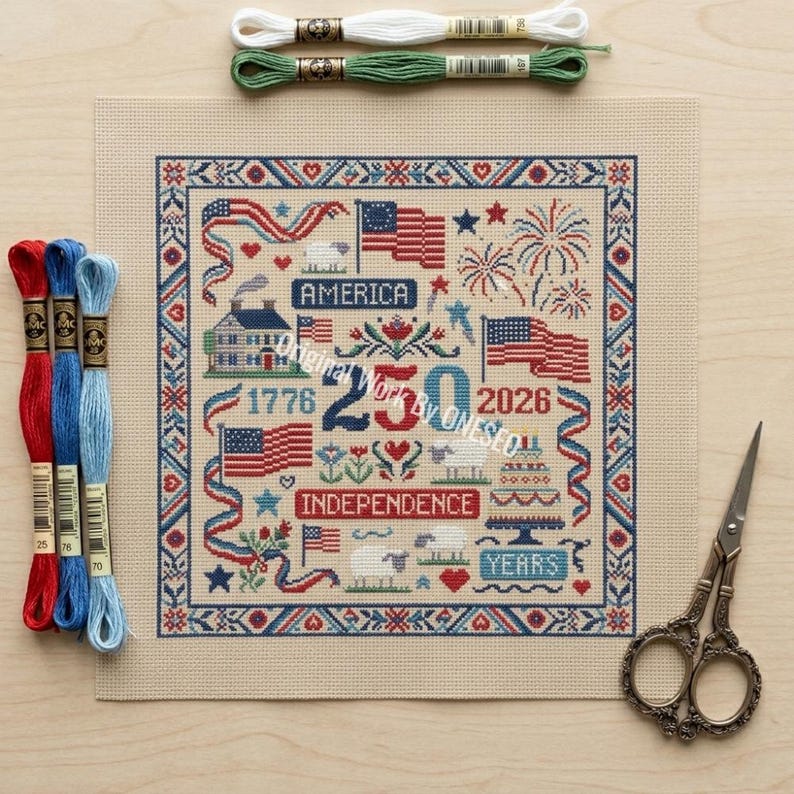 May include: A cross-stitch project with a patriotic theme, featuring American flags, fireworks, and the text "America" and "Independence." The design is framed with a blue and red border. Embroidery floss in red, blue, green, and white, along with scissors, are also visible.