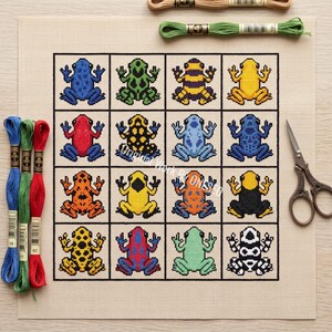 May include: A cross-stitch pattern on beige fabric featuring a grid of sixteen colorful frogs in various patterns and colors. Three skeins of embroidery floss in blue, green, and red are on the left, one green skein is at the top, and metal scissors are on the right.