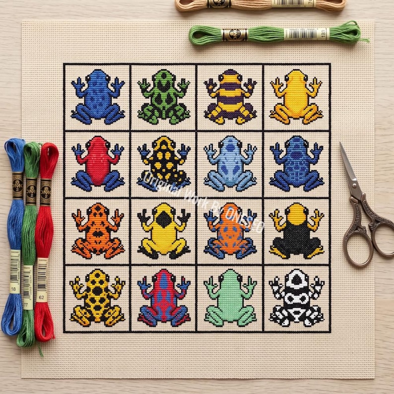 May include: A cross-stitch pattern on beige fabric featuring a grid of sixteen colorful frogs in various patterns and colors. Three skeins of embroidery floss in blue, green, and red are on the left, one green skein is at the top, and metal scissors are on the right.