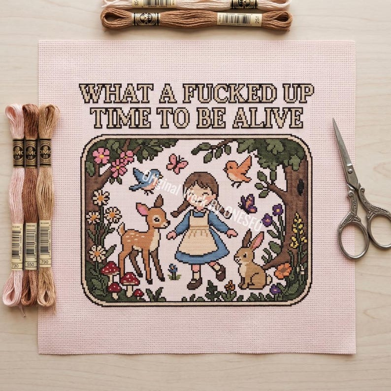 May include: A cross-stitch pattern on light pink fabric featuring a girl in a blue dress, a deer, a rabbit, birds, and flowers within a rectangular border. The text "WHAT A FUCKED UP TIME TO BE ALIVE" is stitched above. Includes embroidery floss and scissors.