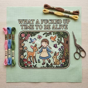May include: A cross-stitch project on light green fabric featuring a person in a blue dress, a deer, a rabbit, birds, and flowers. The text reads "WHAT A FUCKED UP TIME TO BE ALIVE". Includes six embroidery floss skeins in various colors and metal scissors.
