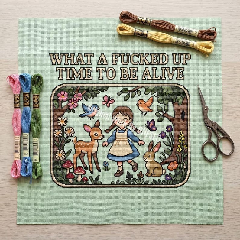 May include: A cross-stitch project on light green fabric featuring a person in a blue dress, a deer, a rabbit, birds, and flowers. The text reads "WHAT A FUCKED UP TIME TO BE ALIVE". Includes six embroidery floss skeins in various colors and metal scissors.