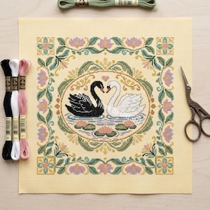 May include: A cross-stitch canvas featuring two swans, one black and one white, facing each other within an oval. The design includes a floral border with pink and green accents. Embroidery floss and scissors are also visible.