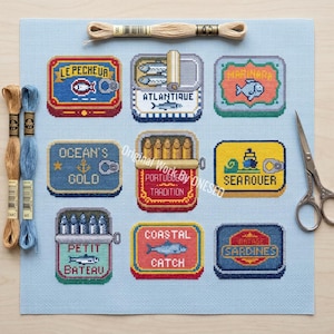 Könnte beinhalten: Kreuzstich-Stickmuster mit neun farbenfrohen Sardinenbüchsen-Designs, darunter Texte wie "OCEAN'S GOLO" und "SEAROUER". Das Set enthält DMC-Stickgarn und eine Schere auf hellblauem Stoff.