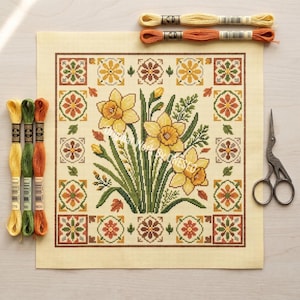 May include: A needlepoint canvas featuring a daffodil floral design with a border of geometric patterns in shades of yellow, orange, and green. Embroidery floss and small scissors are also present.