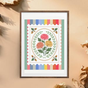 Puede incluir: Arte en punto de cruz enmarcado con un dise&ntilde;o floral. La obra de arte muestra tres crisantemos en rosa, amarillo y melocot&oacute;n, rodeados de hojas verdes y un borde decorativo. El marco es de color marr&oacute;n oscuro.