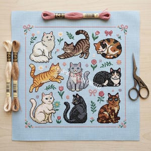 May include: A light blue cross-stitch featuring nine different cat designs, surrounded by floral and bow motifs. The cats are in various poses and colors. Embroidery floss and scissors are also present.