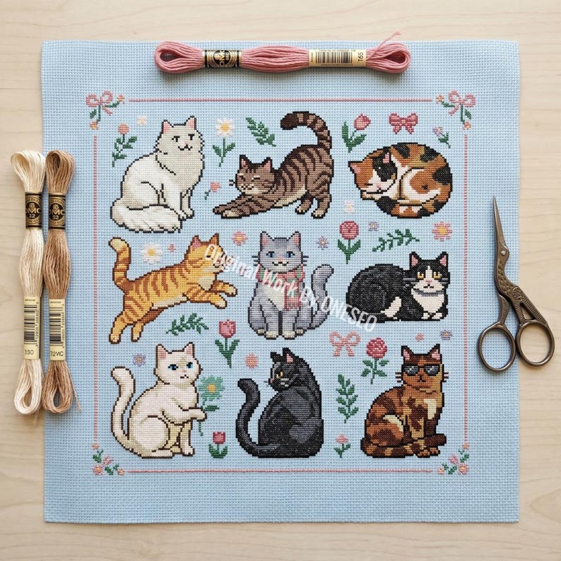 May include: A light blue cross-stitch featuring nine different cat designs, surrounded by floral and bow motifs. The cats are in various poses and colors. Embroidery floss and scissors are also present.