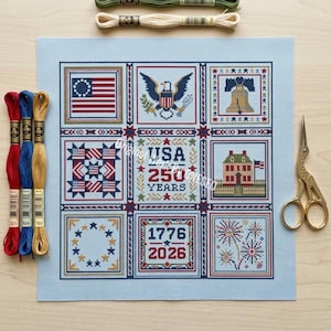 May include: A patriotic needlepoint design featuring American symbols like the flag, eagle, and Liberty Bell. The design includes the text "USA 250 Years" and "1776 2026". Embroidery floss and gold scissors are also visible.