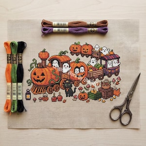 Könnte beinhalten: Ein Kreuzstichmuster auf beigem Stoff mit einem Halloween-Zug aus Kürbissen, Geistern und einer schwarzen Katze. Das Design enthält kleine Kürbisse, Zuckermais und Pilze. Fünf Stickgarnstränge und eine Metallschere sind ebenfalls abgebildet.