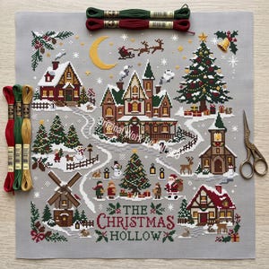 Puede incluir: Una tela de punto de cruz gris con una escena de pueblo navideño con casas, una iglesia, un molino, un gran árbol, Santa Claus y renos. El texto dice THE CHRISTMAS HOLLOW. Incluye tres madejas de hilo de bordar y tijeras de metal.