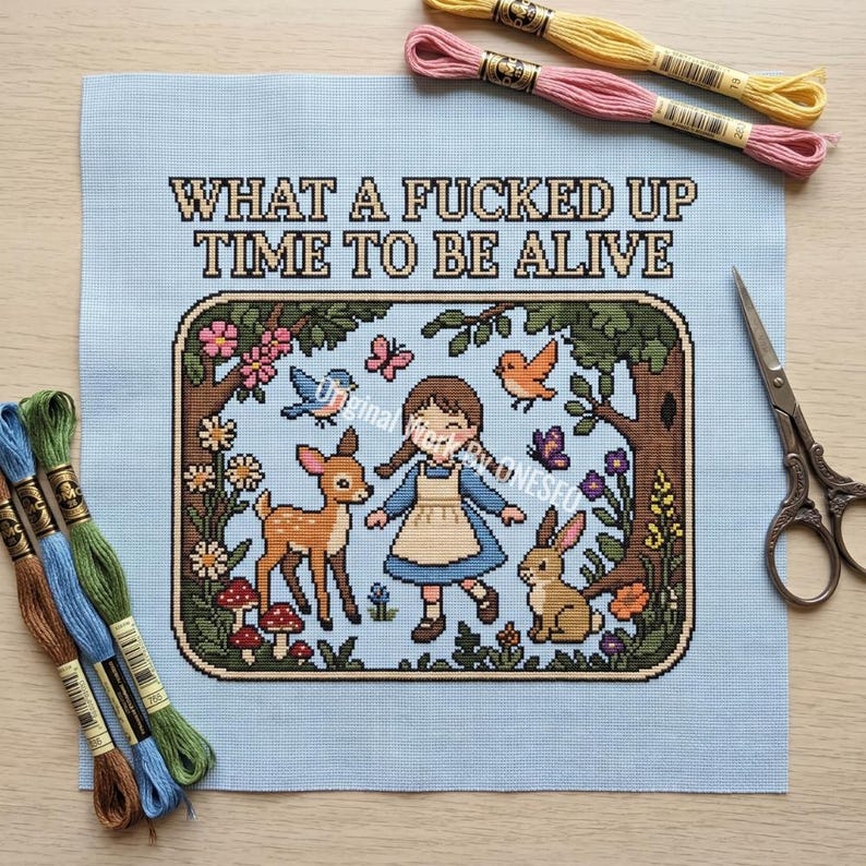 May include: A light blue cross stitch fabric featuring the text WHAT A FUCKED UP TIME TO BE ALIVE above a framed scene of a child, a deer, and a rabbit in a forest. The kit includes embroidery floss in yellow, pink, blue, and green, and metal scissors.