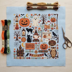 May include: A light blue cross stitch fabric featuring a Halloween-themed pattern with pumpkins, a ghost, black cats, an owl, a haunted house, and text reading Pumpkin Festival, Halloween, and Trick or Treat, accompanied by embroidery floss and scissors.