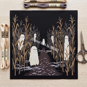 May include: A cross-stitch embroidery on black fabric depicting a path through a cornfield with several white ghost figures. The design includes brown and tan corn stalks and a dark path. Embroidery floss skeins and metal scissors are nearby.