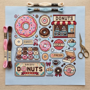 Peut inclure: Un tissu de point de croix bleu clair avec un thème de boutique de beignets, incluant des illustrations de beignets, une devanture de boulangerie, du café et le texte DONUTS, BAKERY, SWEET et FRESH. Avec quatre échevettes et des ciseaux.