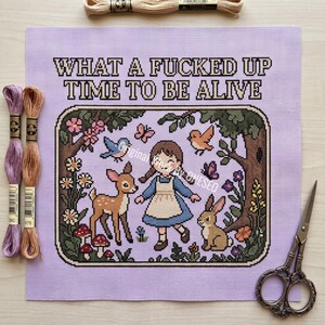 May include: A cross-stitch project on light purple fabric featuring a girl, a deer, and a rabbit in a forest scene with the text WHAT A FUCKED UP TIME TO BE ALIVE. Includes two skeins of embroidery floss and a pair of ornate metal scissors.