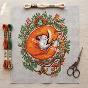 May include: Cross-stitch embroidery of a sleeping fox embracing a white rabbit, surrounded by a wreath of greenery, mushrooms, and a bird. Includes embroidery floss, scissors, and the text "Original Work By ONESEO".