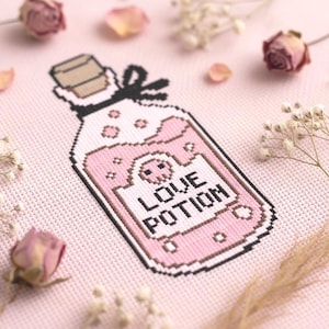 Könnte beinhalten: Ein Kreuzstich-Design einer rosa Zaubertrankflasche mit den Worten "LOVE POTION" auf der Vorderseite. Die Flasche hat ein Totenkopf-Design und ist von getrockneten Blumen und Blütenblättern auf rosa Hintergrund umgeben.