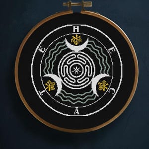 May include: A black cross-stitch embroidery hoop featuring a white and yellow design with the letters H, E, A, and a symbol. The design includes crescent moons, a maze, and wavy lines. The hoop is framed in a wooden ring.