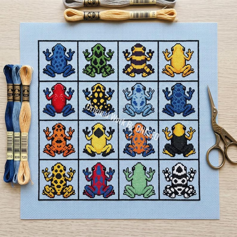 May include: A cross-stitch pattern on light blue fabric featuring a grid of sixteen colorful poison dart frogs in various patterns and colors. Three skeins of embroidery floss in blue and tan shades are placed on the left, with two more at the top.