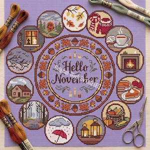 May include: A cross-stitch design on a lavender background, featuring a central circle with "Hello November" and autumn-themed illustrations. The design includes images of leaves, a cozy scarf, a teacup, a fireplace, and a pumpkin pie.