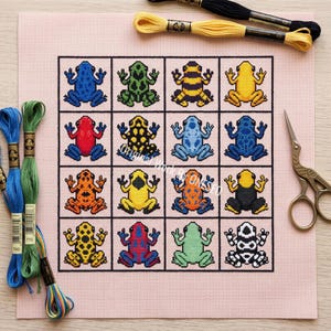 May include: A cross-stitch pattern on light pink fabric featuring a grid of sixteen colorful frogs in various patterns and colors. The design includes blue, green, yellow, red, orange, and black frogs. Embroidery floss and metal scissors are included.