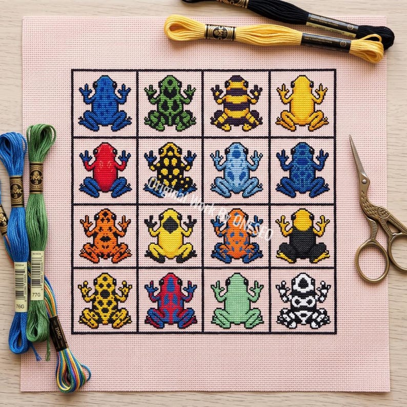 May include: A cross-stitch pattern on light pink fabric featuring a grid of sixteen colorful frogs in various patterns and colors. The design includes blue, green, yellow, red, orange, and black frogs. Embroidery floss and metal scissors are included.