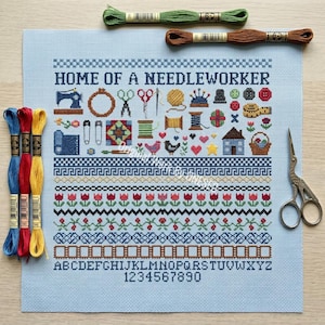 May include: A light blue cross-stitch fabric featuring the text HOME OF A NEEDLEWORKER, various sewing-themed icons, decorative borders, the alphabet, and numbers. Three skeins of embroidery floss in blue, red, and yellow, and metal scissors.