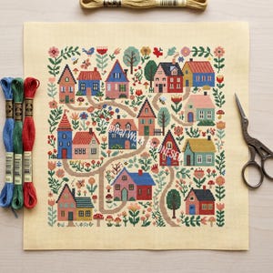 May include: A cross-stitch project featuring a colorful village scene with houses, trees, and flowers. The design is on a cream-colored canvas, with embroidery floss in blue, green, and red. A pair of scissors and a spool of thread are also visible.
