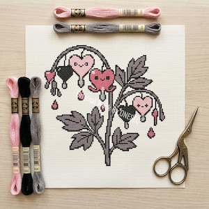 Peut inclure: Un projet de point de croix représentant un cœur saignant stylisé avec des fleurs en forme de cœur roses et noires et des feuilles grises. Des fils à broder roses, noirs et gris et de petits ciseaux sont disposés autour de la toile. La toile est sur une surface en bois clair.