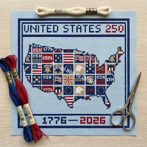 May include: A light blue needlepoint canvas featuring a map of the United States, with each state represented by a flag or emblem. The text "UNITED STATES 250" and "1776-2026" are displayed. Embroidery floss and scissors are also present.