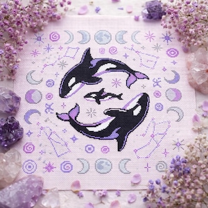 Peut inclure: Une broderie au point de croix représentant trois orques noires et blanches, entourées d'éléments célestes violets : lunes, étoiles et constellations. Le fond est en tissu rose clair. L'image est encadrée de fleurs et de cristaux violets.