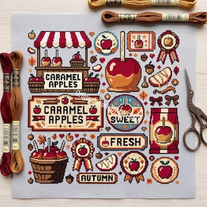 May include: A cross-stitch pattern on light gray fabric featuring various caramel apple motifs, including a stand, baskets, ribbons, and text reading CARAMEL APPLES, SWEET, FRESH, and AUTUMN. Includes two skeins of embroidery floss and metal scissors.