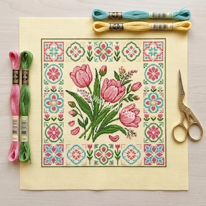 May include: A needlepoint canvas with a floral design of pink tulips and green leaves, framed by a decorative border in pink, teal, and green. Includes embroidery threads in pink, green, teal, and yellow, plus gold scissors.