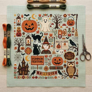 May include: A light green cross-stitch fabric featuring a Halloween sampler with pumpkins, a ghost, black cats, an owl, a haunted house, and trees. Text includes "PUMPKIN FESTIVAL", "HALLOWEEN", and "TRICK OR TREAT". Two embroidery floss skeins and scissors.