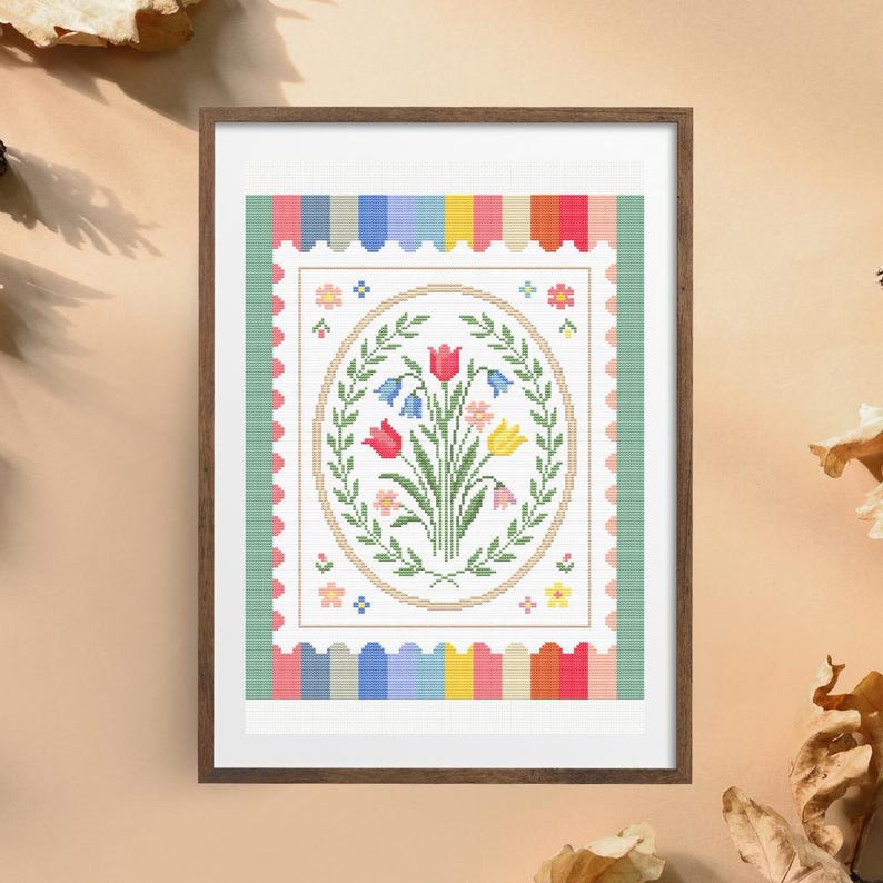 Puede incluir: Una obra de arte en punto de cruz enmarcada con un ramo floral en un marco ovalado. El ramo incluye tulipanes rojos, campanillas azules y flores amarillas. La obra est&aacute; rodeada por un borde a rayas de colores y un borde festoneado.