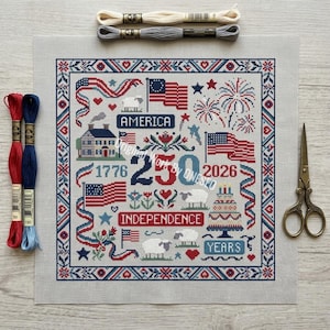 May include: A cross-stitch design with patriotic themes, including flags, fireworks, and the text "America" and "Independence". The design is framed with red and blue borders and includes the years 1776 and 2026. Embroidery floss and scissors are also present.