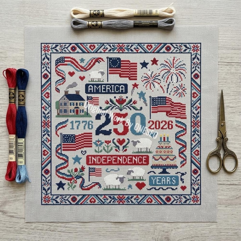 May include: A cross-stitch design with patriotic themes, including flags, fireworks, and the text "America" and "Independence". The design is framed with red and blue borders and includes the years 1776 and 2026. Embroidery floss and scissors are also present.