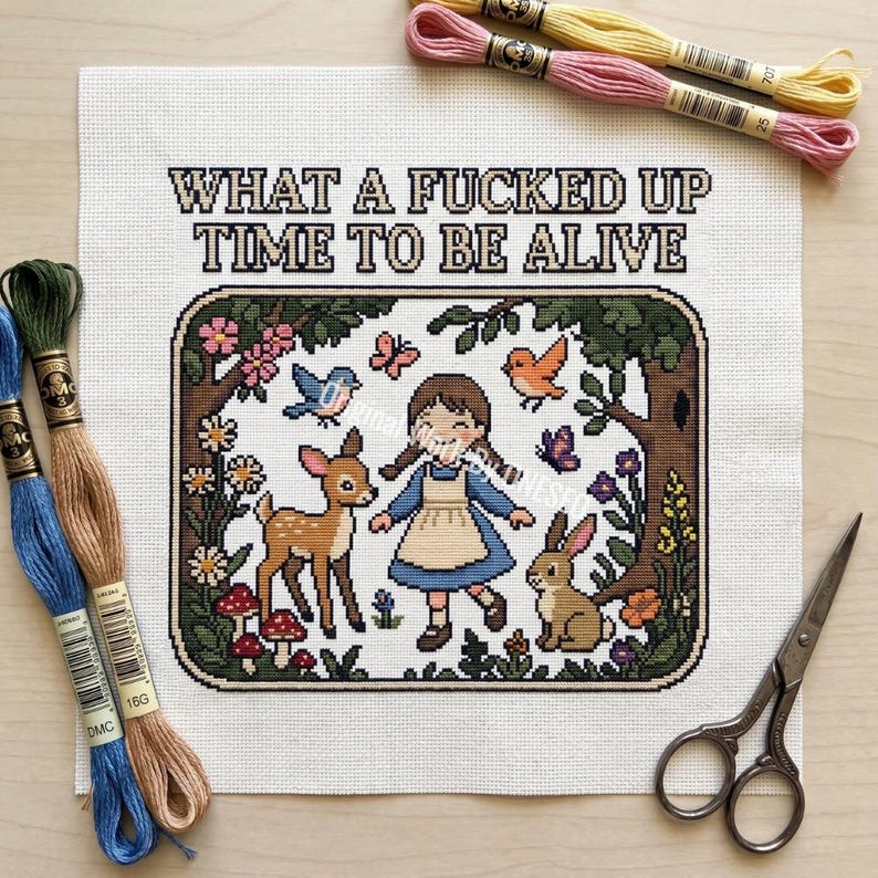 May include: A cross-stitch pattern on white fabric featuring a girl, a deer, and a rabbit in a forest, with the text "WHAT A FUCKED UP TIME TO BE ALIVE" above. Includes embroidery floss in blue, green, tan, pink, and yellow, and metal scissors.