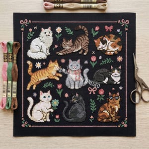 May include: A black needlepoint featuring nine different cat designs in various poses, surrounded by floral and bow accents. The cats are in shades of white, gray, orange, brown, and black. Embroidery floss and scissors are visible.