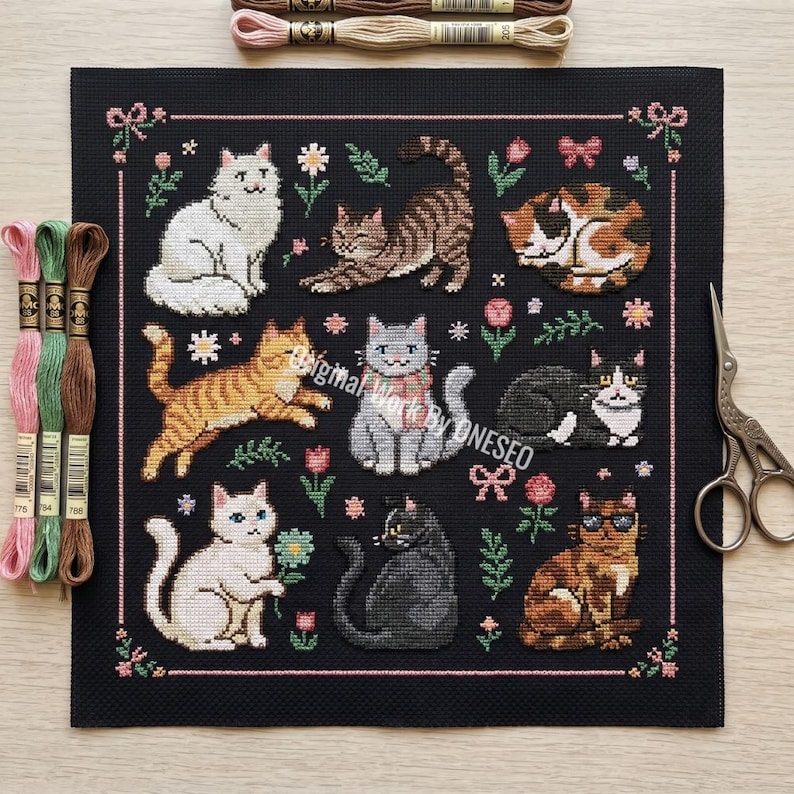 May include: A black needlepoint featuring nine different cat designs in various poses, surrounded by floral and bow accents. The cats are in shades of white, gray, orange, brown, and black. Embroidery floss and scissors are visible.