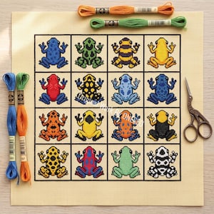 Peut inclure: Un motif de point de croix sur tissu beige présentant une grille de seize grenouilles colorées aux motifs variés. L'ensemble comprend des échevettes de fil à broder orange, vert, bleu, jaune, rouge et noir ainsi qu'une paire de ciseaux.