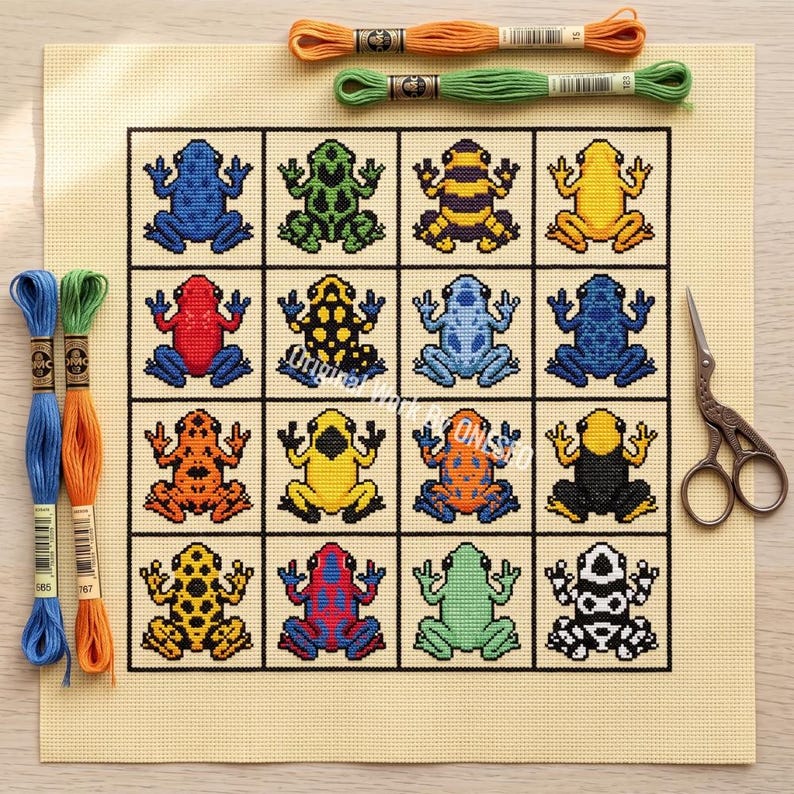May include: A cross-stitch pattern on beige fabric featuring a grid of sixteen colorful frogs in various patterns and colors. The set includes orange, green, blue, yellow, red, and black embroidery floss skeins and a pair of metal scissors.
