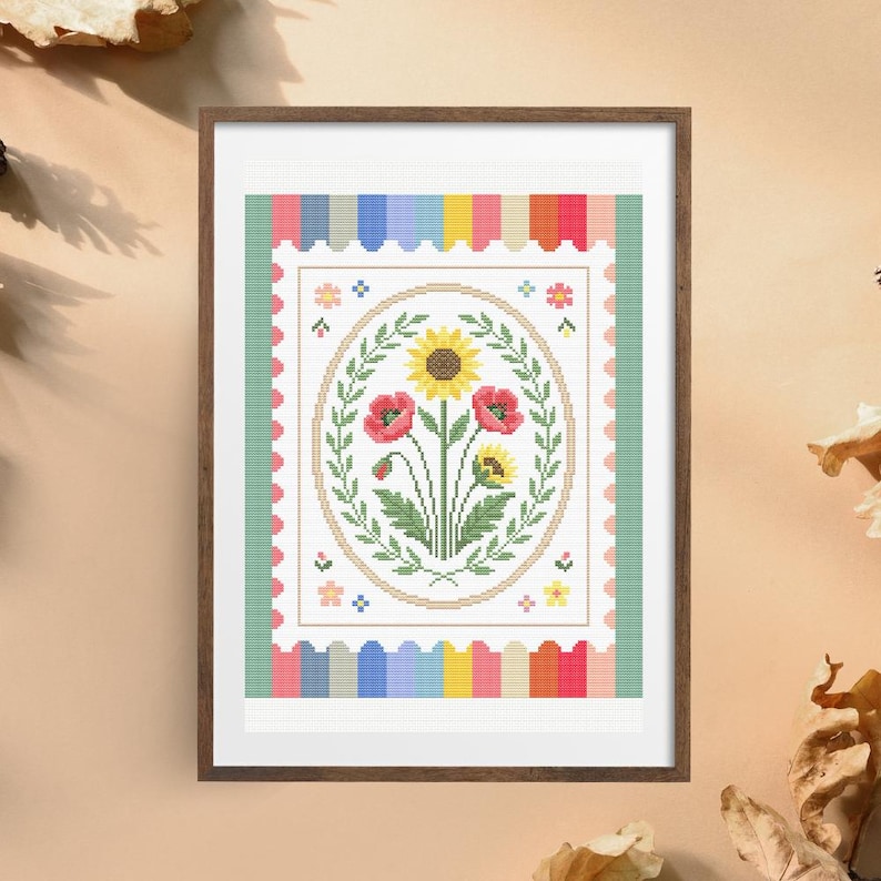 Puede incluir: Arte de punto de cruz enmarcado con un dise&ntilde;o floral. La obra de arte muestra un &oacute;valo central con girasoles y amapolas rojas, rodeado de hojas verdes. El marco tiene un borde a rayas de colores y un marco de madera.