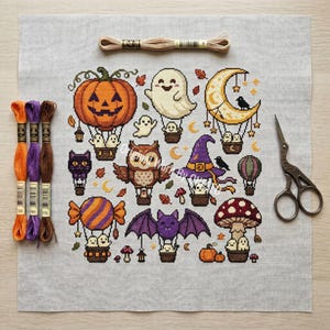 May include: A cross-stitch pattern on light gray fabric featuring Halloween motifs including hot air balloons with ghosts, a pumpkin, an owl, a bat, a crescent moon, and mushrooms. Three embroidery floss skeins and vintage-style scissors are included.