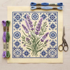 May include: A needlepoint canvas with a lavender bouquet and a blue tile border. Embroidery floss in purple, green, and blue, plus vintage scissors, surround the canvas. The canvas measures approximately 25 cm by 25 cm.