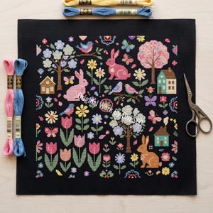 May include: A black needlepoint canvas featuring a colorful spring design with pink rabbits, birds, flowers, trees, and houses. The canvas is surrounded by embroidery floss in shades of pink, blue, and yellow, along with a pair of scissors.